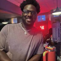 Man smiling at Do it a-Ginn Event Hall, lively atmosphere, vibrant red lighting, casual celebration, people enjoying event, social gathering, entertainment venue, happy guest, modern event space, nightlife scene, stylish decor, community event, celebration ambiance, indoor party setting, fun night out, young adult, entertainment venue, live event, event hall ambiance, youthful energy.