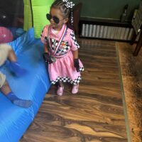 Cute girl dressed as a race car driver at Do it a-Ginn Event Hall, enjoying a children's party with fun activities like bounce houses, wearing racing-themed costume and accessories.